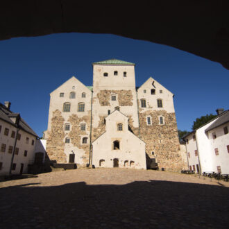 Turku Castle’s stone keep and white-walled buildings rise around a cobbled courtyard, framed by a shadowy stone arch under a deep blue sky.