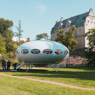 Visitors stand beside a pale white Futuro house at the Kaisaniemi Botanic Garden in Helsinki, where the UFO-shaped structure with oval windows sits on metal legs against a backdrop of trees and a large historic building.