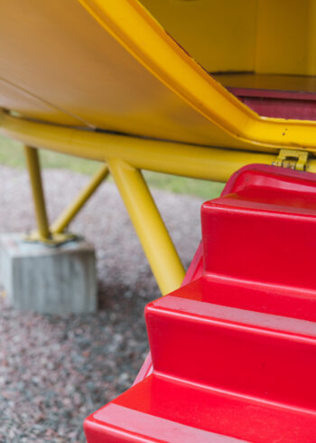 Bright red folding steps lead up to a yellow Futuro house, with yellow support struts and a metal foot visible beneath the structure.