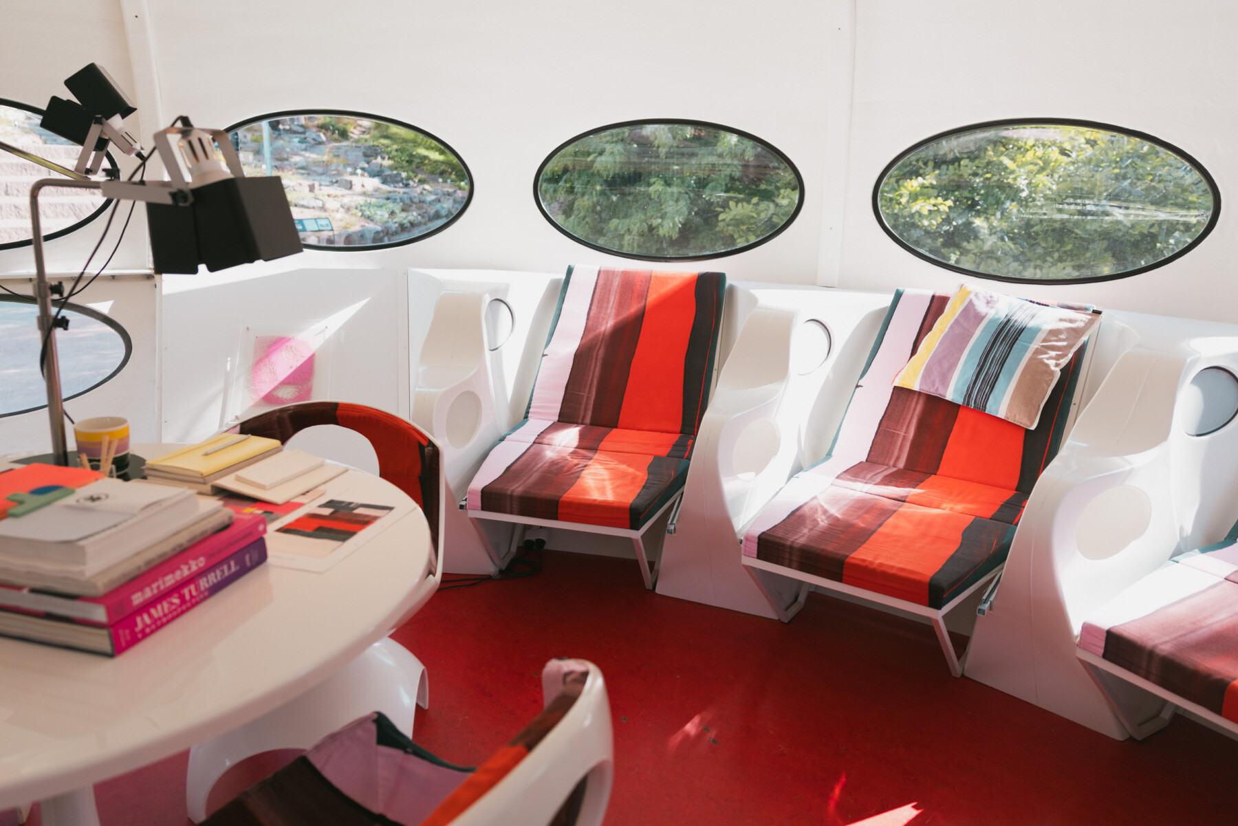 The Futuro’s bright interior has a red floor, curved white wall panels and three large oval windows showing trees and buildings outside, while sculptural white chairs are upholstered in striped red Marimekko textiles.