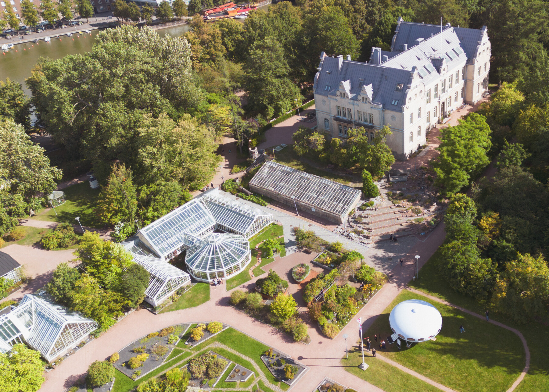 An aerial view of the Kaisaniemi Botanic Garden shows its glasshouses and landscaped gardens among dense trees, with a large historic building and the white UFO-shaped Futuro positioned on the open lawn.