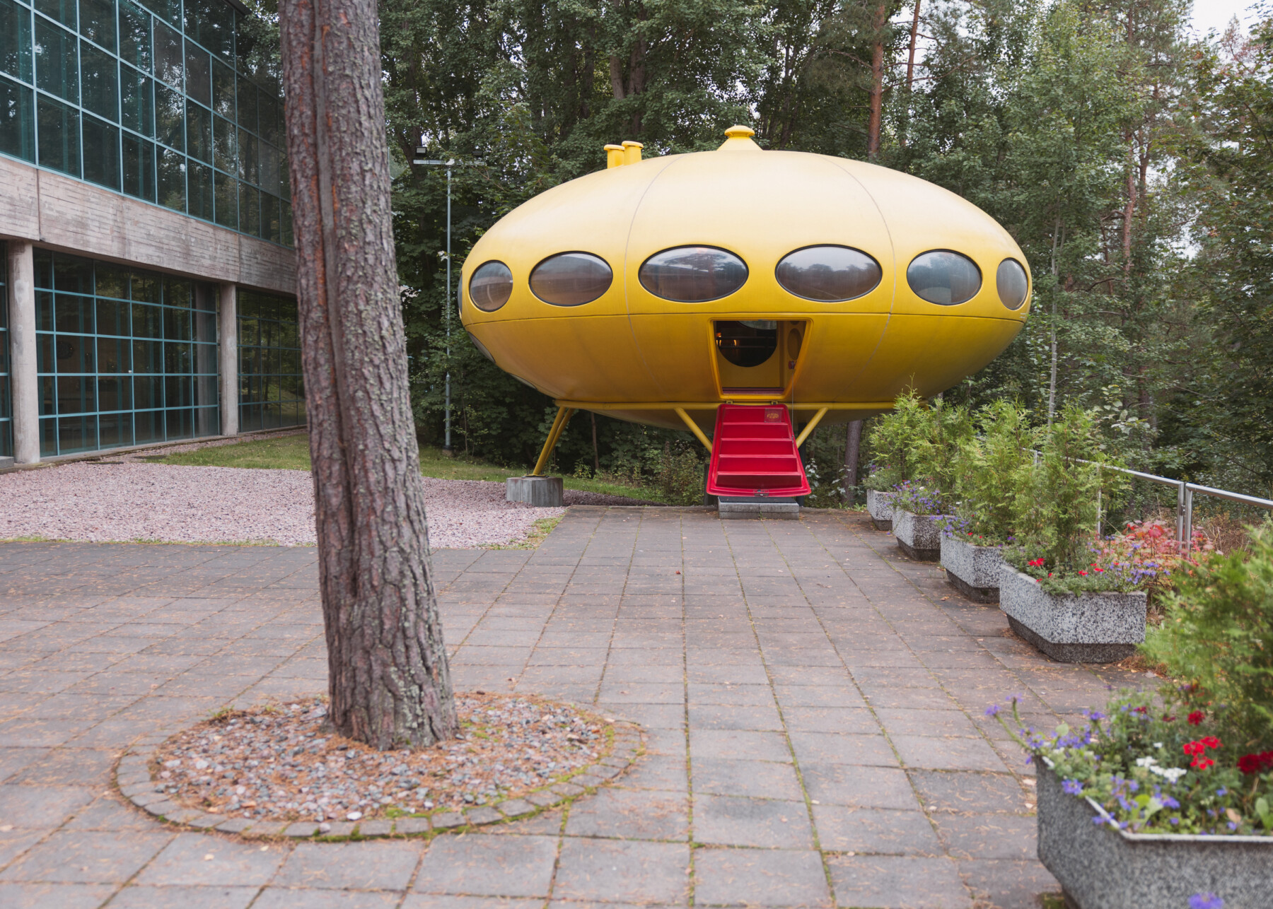 A yellow Futuro No. 001 stands on supports behind EMMA – Espoo Museum of Modern Art – with oval windows and a red staircase leading to its entrance.