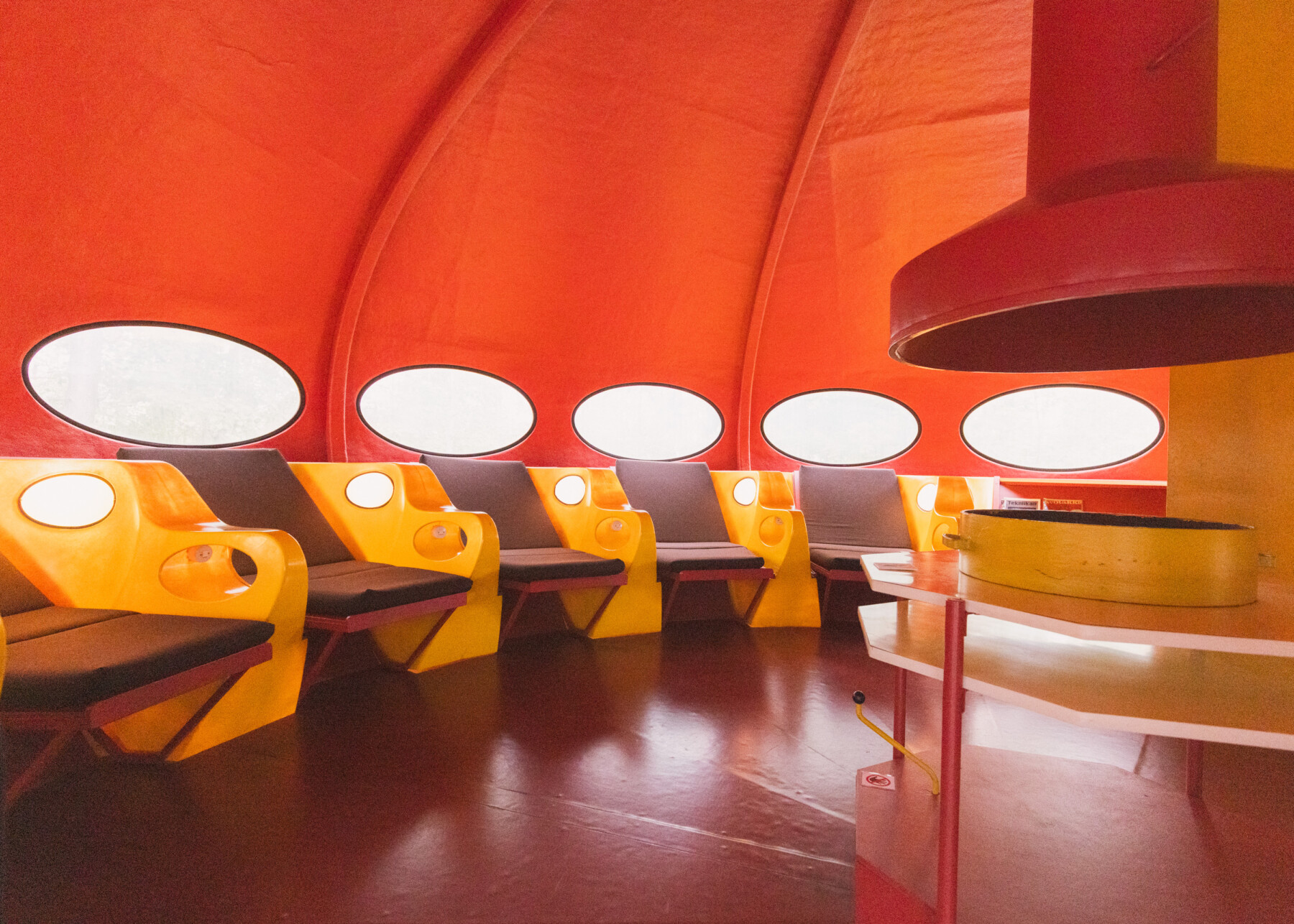 Bright yellow moulded seats with oval white panels stand against the curved red-orange walls of a Futuro interior, with a polished dark floor and a row of oval windows.