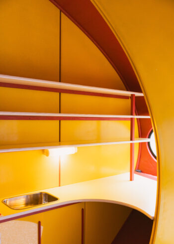 The Futuro’s compact interior features a curved kitchen counter with a stainless-steel sink, yellow walls, red structural details and a round window.