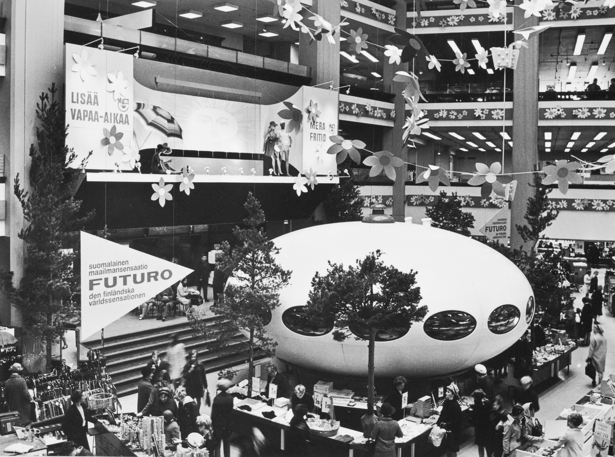 A Futuro house with dark oval windows stands on display at Helsinki’s Stockmann department store, surrounded by small trees, flower decorations and crowds of shoppers.