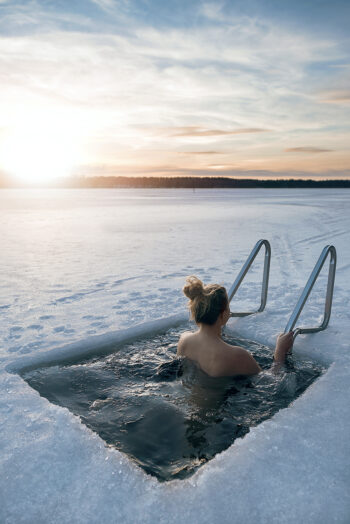 Uma mulher está imersa até os ombros em água em um buraco retangular no gelo de um lago congelado, enquanto se segura em uma escada que está na borda do buraco.