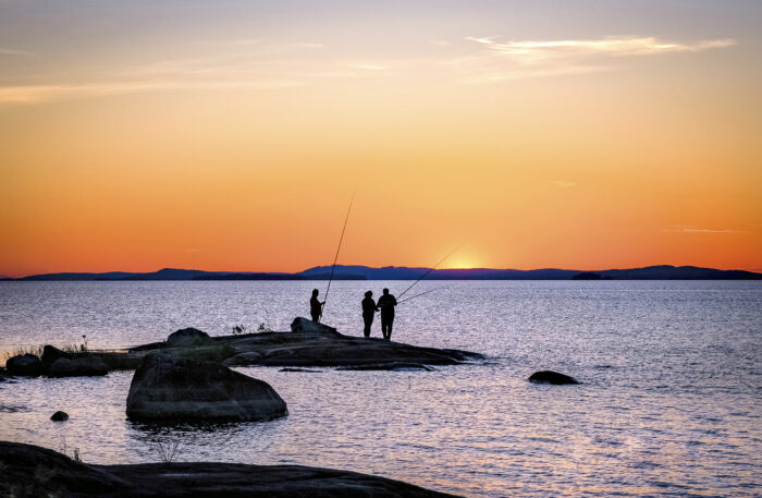 Drei Menschen mit Angelruten stehen auf einem großen Felsen am Ufer.