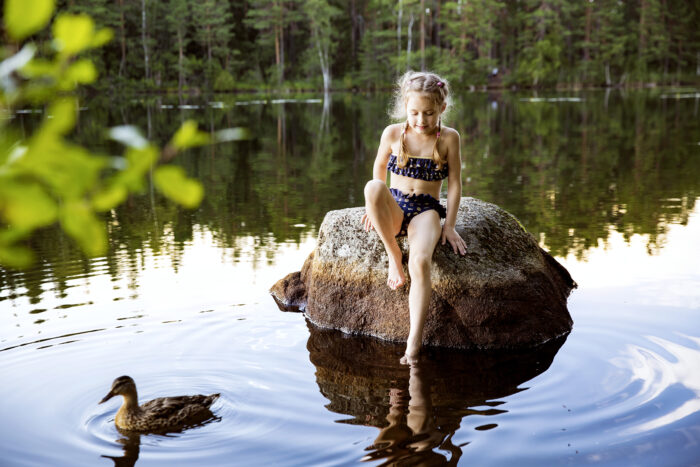 Sentada sobre una roca en un lago, una niña en bañador introduce la punta del pie en el agua, mientras un pato pasa nadando a su lado.