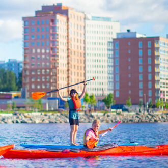 Duas pessoas em caiaques e uma pessoa em uma prancha de stand-up paddle acenam para a câmera de um lago, com prédios de apartamentos ao fundo.
