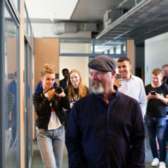 A man wearing a beret and a grey goatee walks down a hall at a university building with students who are taking pictures with cameras and phones.
