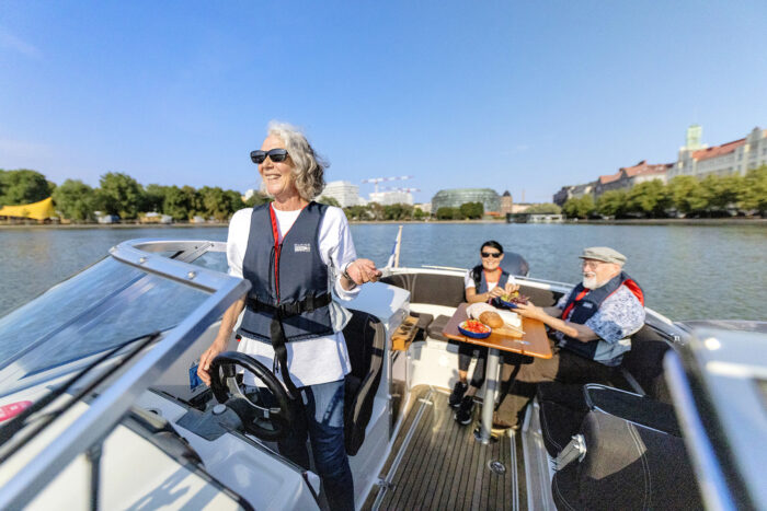 Drei lächelnde Menschen sitzen in einem Motorboot vor der Küste Helsinkis, einer von ihnen fährt, und die anderen beiden essen.